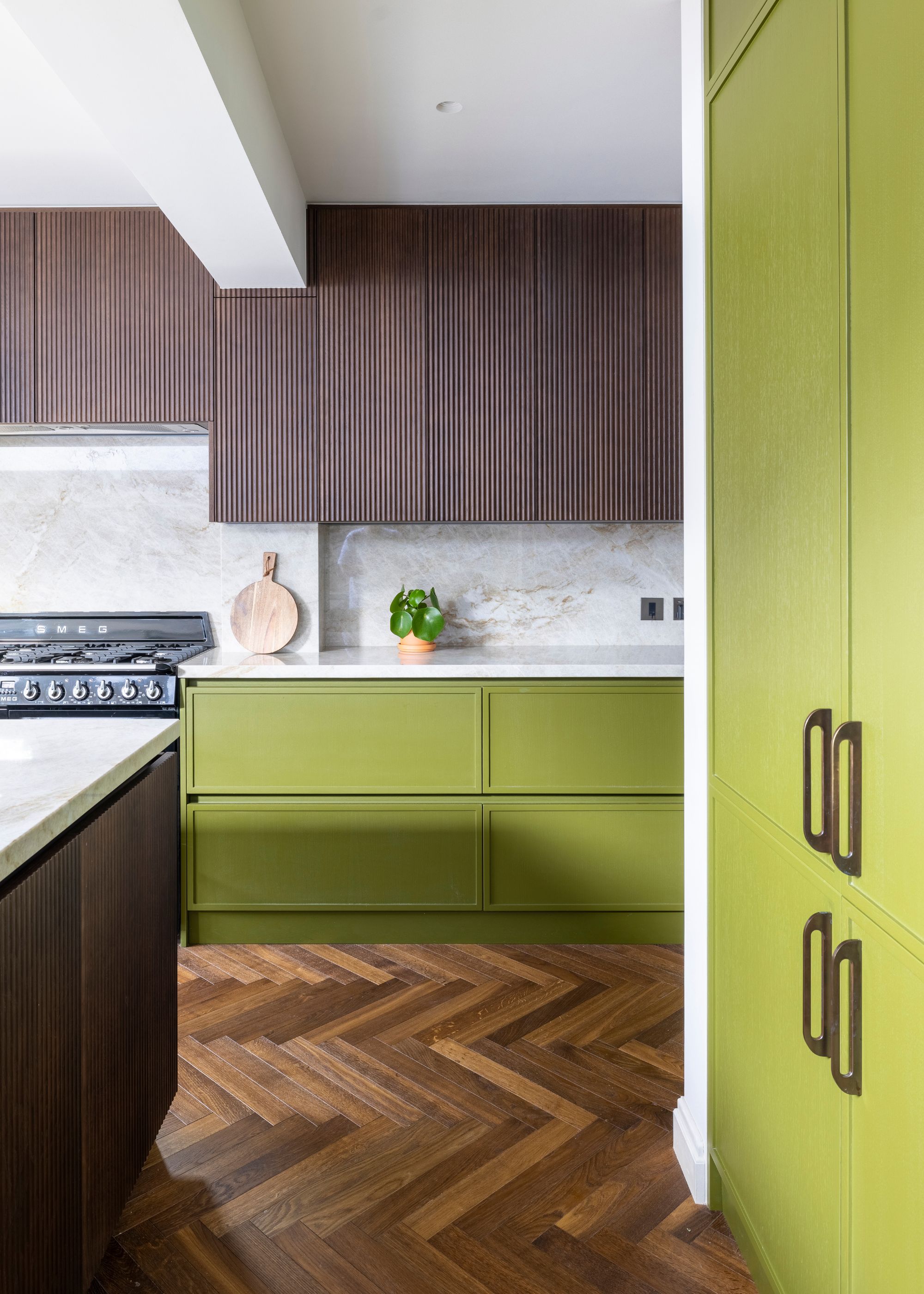 A sleek modern kitchen with olive green lower cabinets and pantry, dark wood upper cabinets and island, white stone countertops and backsplash, and parquet flooring.