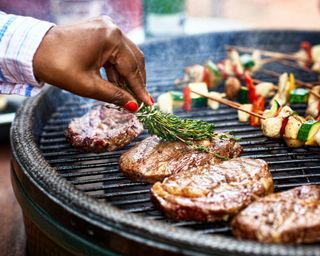 Steaks being seasoned on an outdoor BBQ grill with vegetables and peppers behind