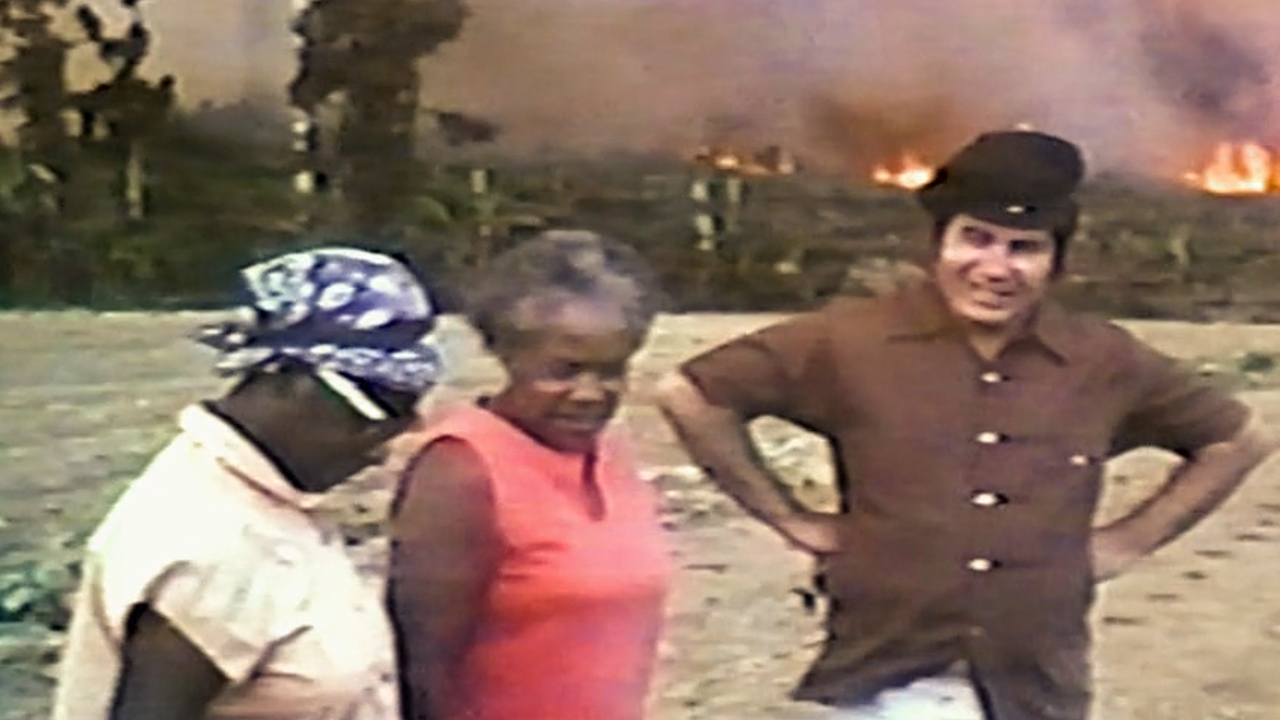 Jim Jones in a brown shirt and beret, talking to some of the church members in archival footage