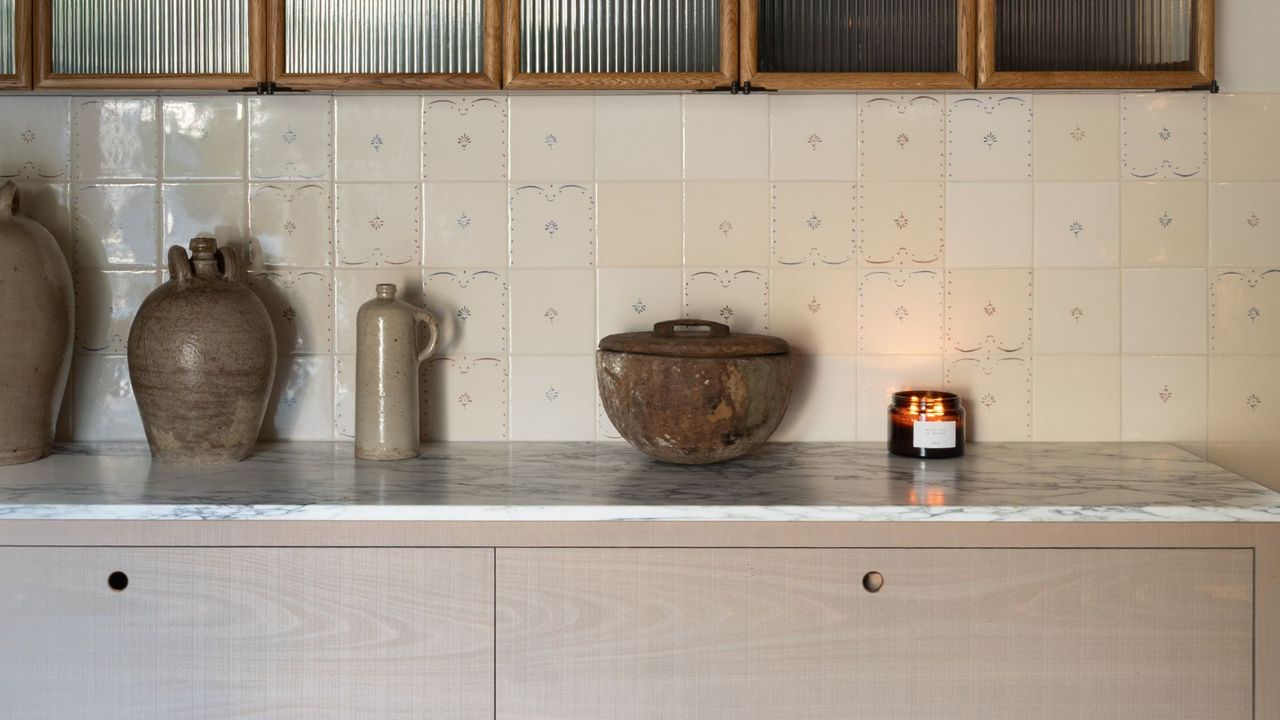 Small kitchen countertop in marble, with deVOL Kitchen Ditsy Delft wall tiles. These are small, varied shades of cream with tiny floral design in center of some of the tiles, not all. Ceramic pots can be seen on the counter, as well as the top drawers