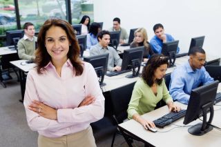 Female teacher stands in front of students using desktop computers.