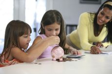 Two sisters inserting coins into a piggy bank with their mother smiling near them