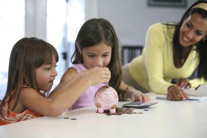 Two sisters inserting coins into a piggy bank with their mother smiling near them