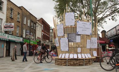 View of Union Street and a tall, circular light wood panel structure with multiple pieces of paper in different sizes with print attached to it. There are shops with flats above, a person on a bike and a number of other people in the background