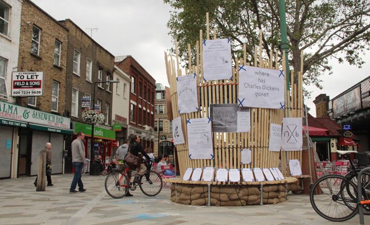 View of Union Street and a tall, circular light wood panel structure with multiple pieces of paper in different sizes with print attached to it. There are shops with flats above, a person on a bike and a number of other people in the background