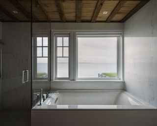 A bathroom with sea views from oversized window, white bath and wooden beamed ceiling