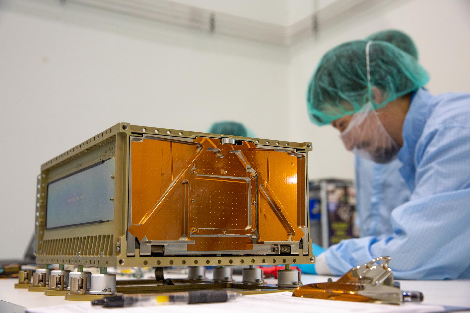 NASA scientists in clean suits work on a small shoebox-sized satellite.