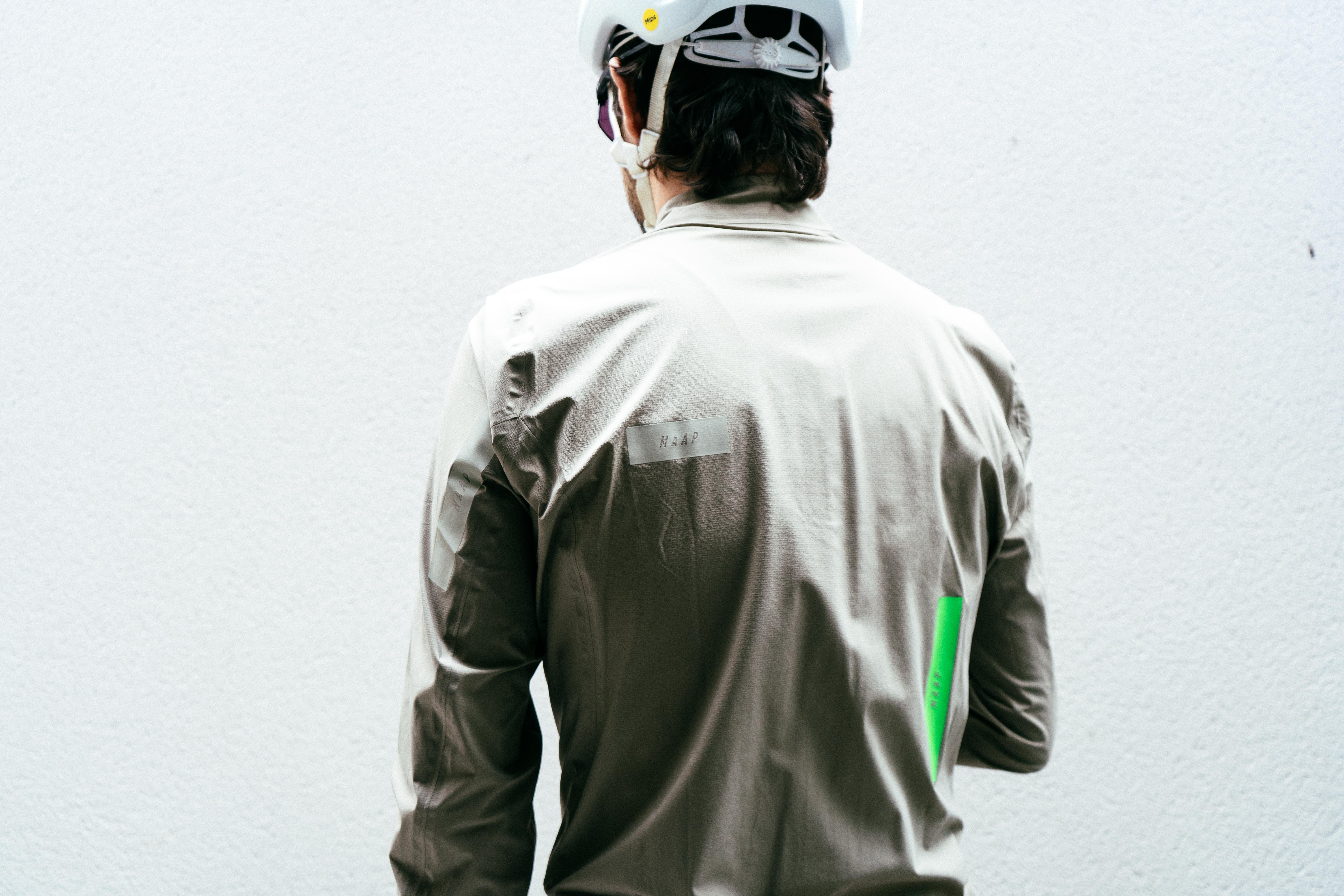 A man in an off-white road cycling rain jacket faces a white wall