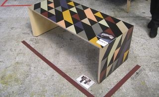 Grey floor, red tape line, multi-coloured triangular shape pattern coffee table, persons feet in black shoes and black trousers at the top right of the shot