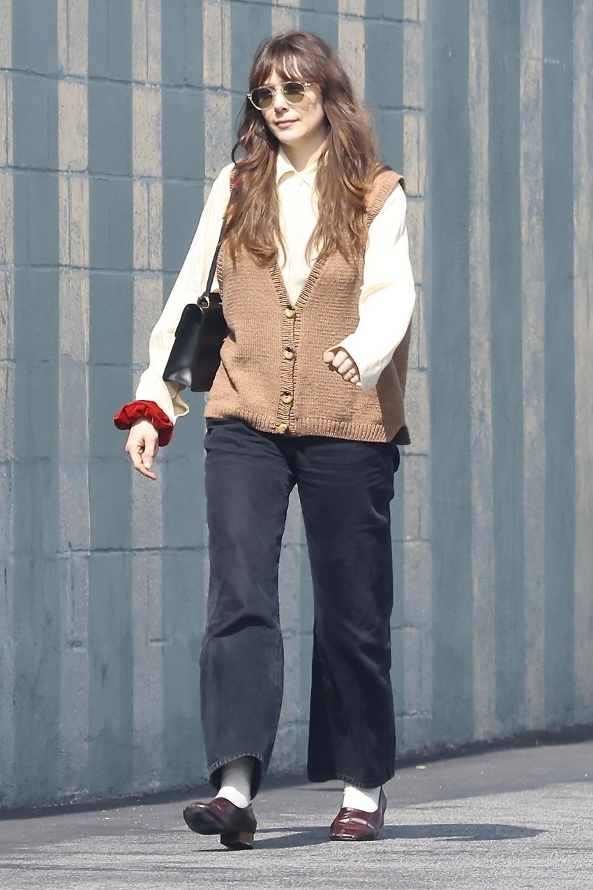 Elizabeth Olsen wearing a tan sweater vest, button-down shirt, jeans, and high-vamp pumps.