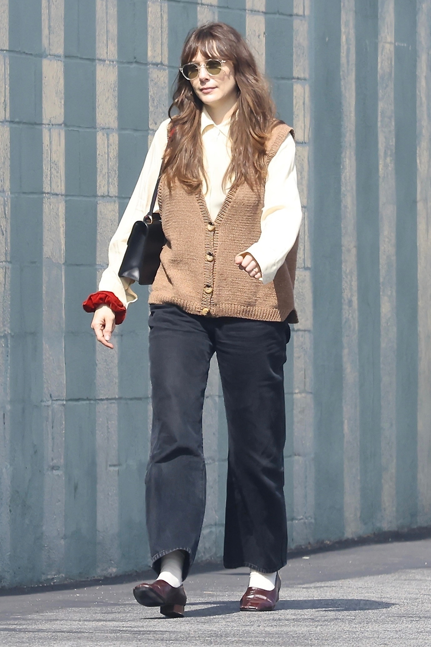 Elizabeth Olsen wearing a tan sweater vest, button-down shirt, jeans, and high-vamp pumps