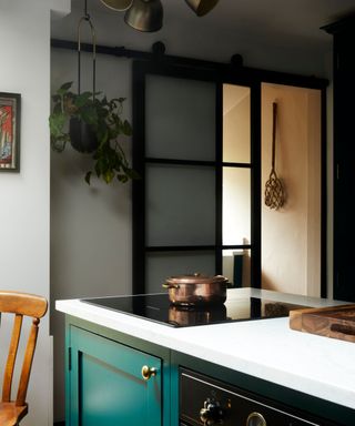 A kitchen island with a white surface, black hob with copper pot, and dark green cabinets, with a black sliding door behind it