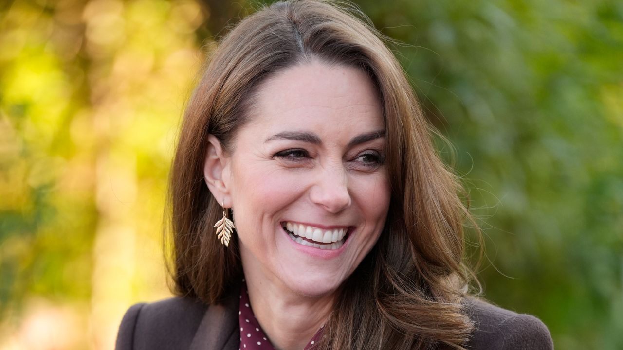 Kate Middleton smiling and wearing a brown coat with gold leaf earrings