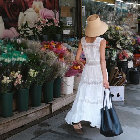 Shay wears a straw hat, white lace-trim maxi dress, and black leather bag.