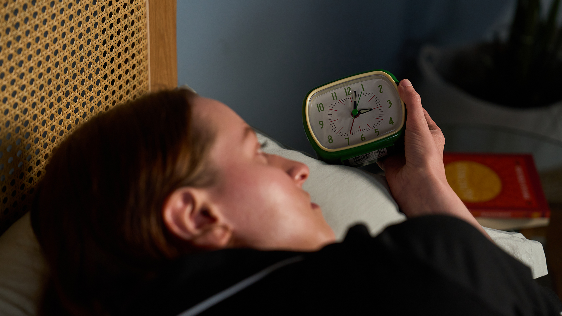 A woman with long dark brown hair lies in bed holding a green alarm clock that reads 3 a.m.