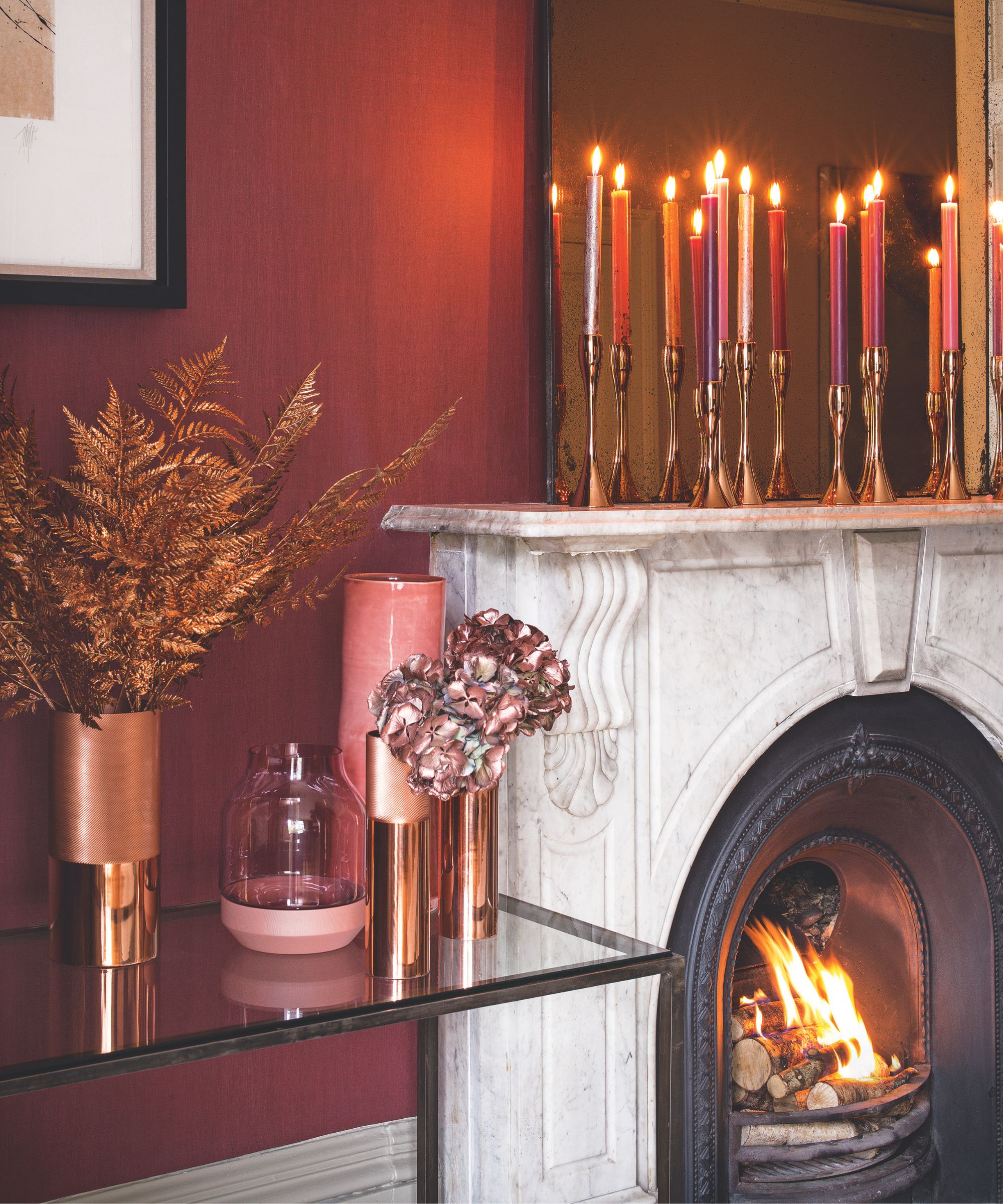 A marble mantelpiece with a lit fire in the fireplace, with gold candle holders and colorful taper candles on the mantelpiece, and a glass table to the left with gold dried flowers and pink vases. The walls are deep red.