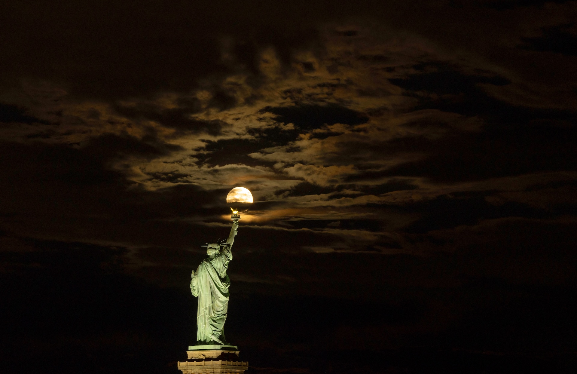 The moon rises behind the Statue of Liberty, making it look like the statue is holding the moon