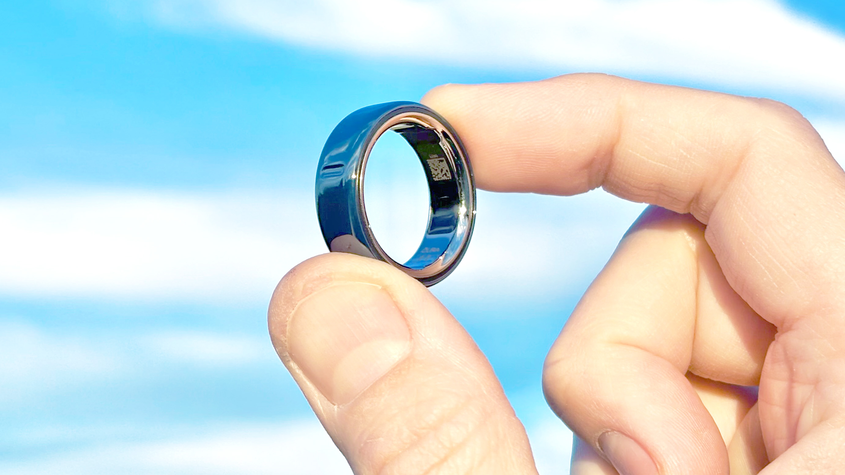 Close-up of the Oura Ring 4 Ceramic shining in the sunlight in a user's hand with blue skies and clouds behind it