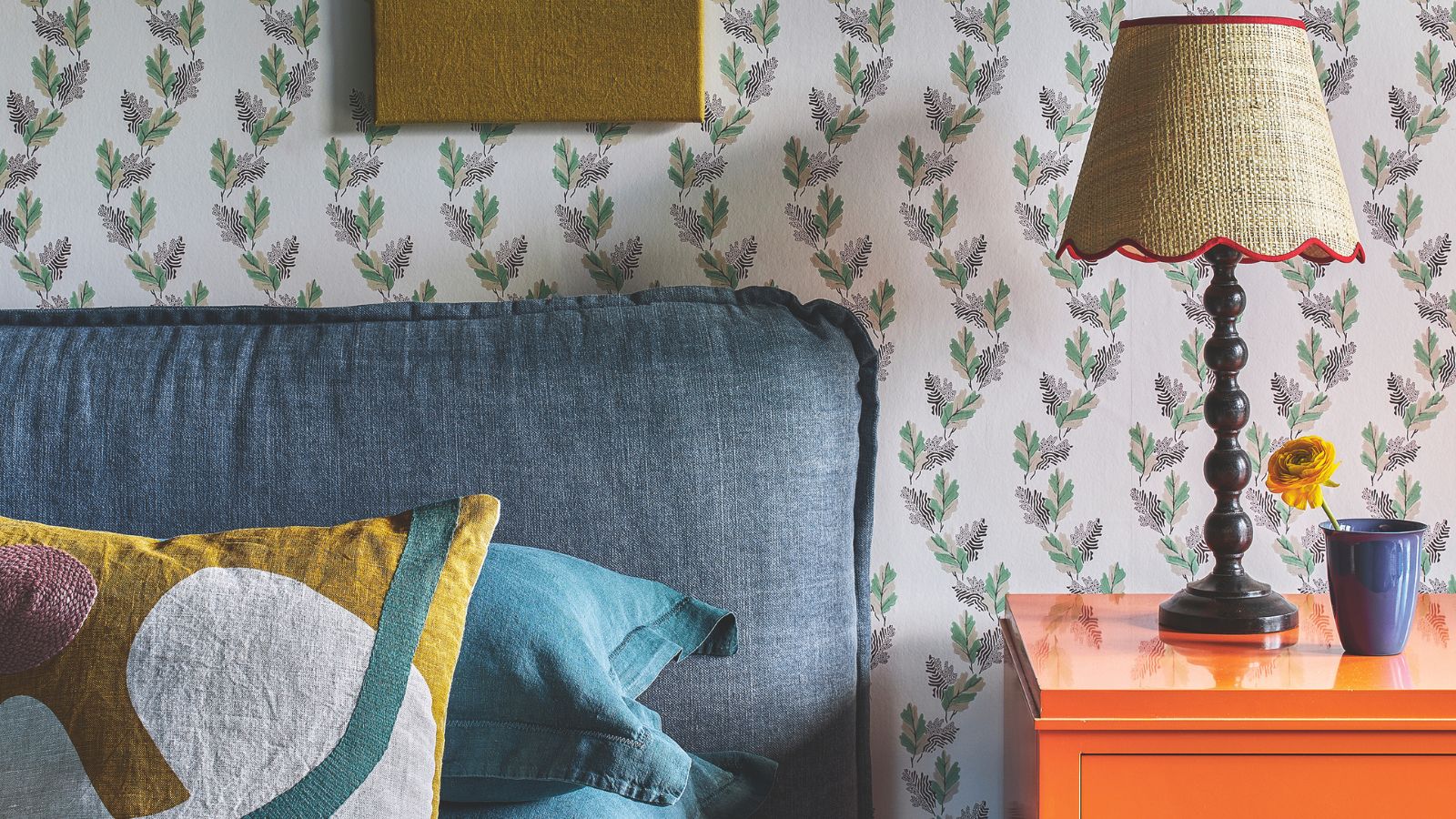 Blue headboard with patterned cushions, beside an orange bedside table with lamp with rattan, scalloped shade and blue vase with a flower. Behind is floral patterned wallpaper.