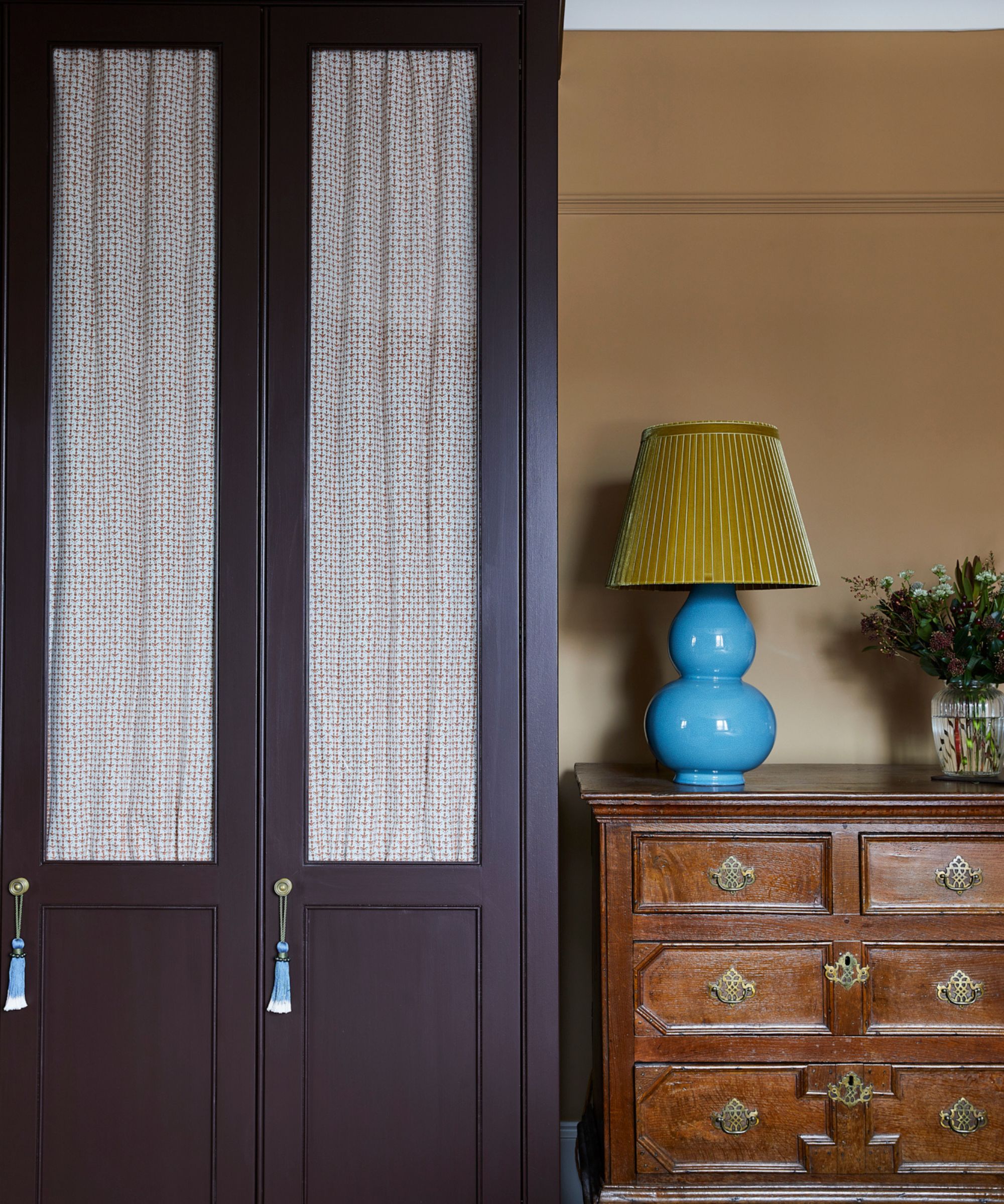 Bedroom with dark purple painted wardrobe with curtained panels, a vintage wooden dresser with a lamp with a turquoise base and yellow shade on top, and a vase of flowers.