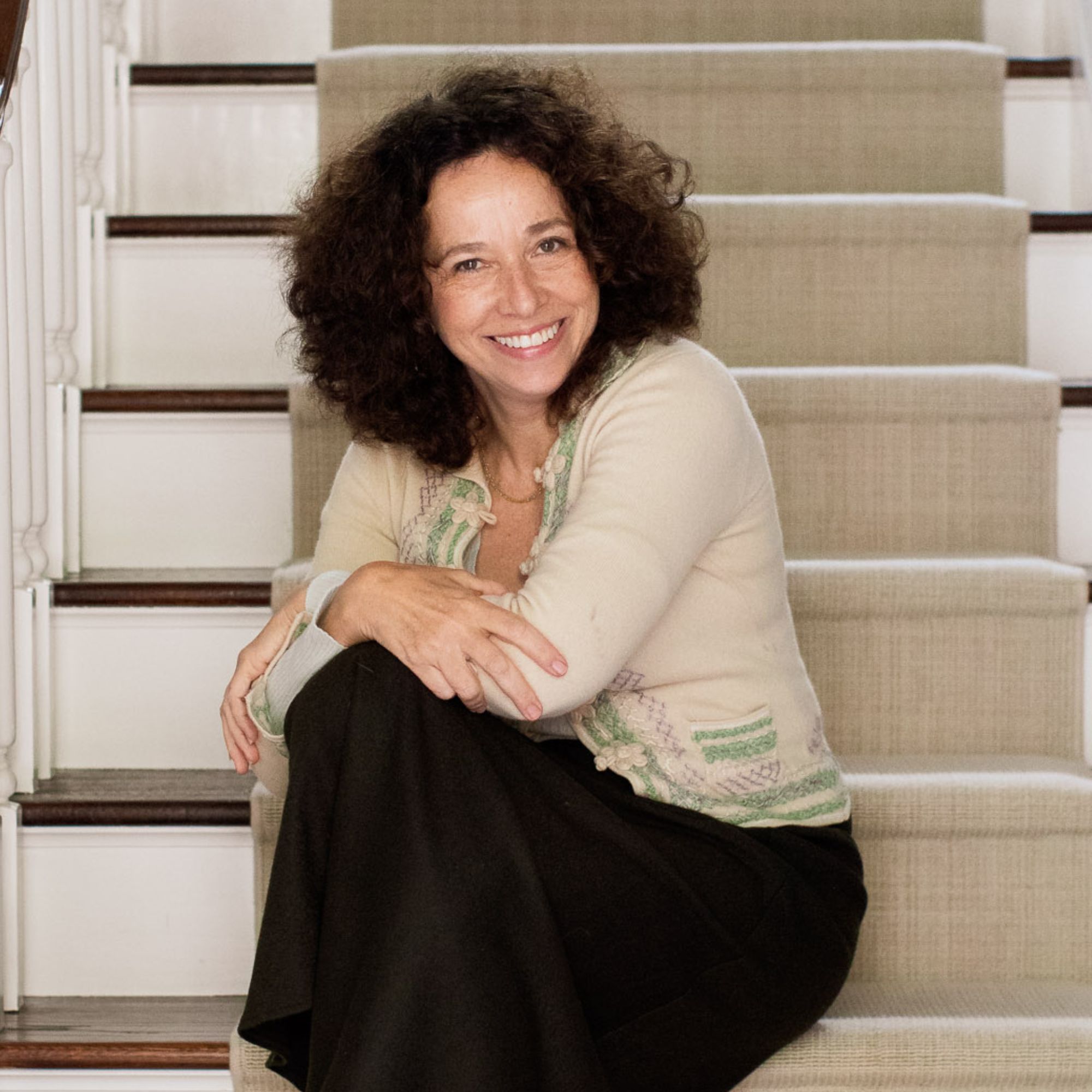 A picture of Amalia Gal, a woman with dark brown frizzy hair wearing a beige top and black skirt sitting on white steps with a beige runner rug in the middle of it