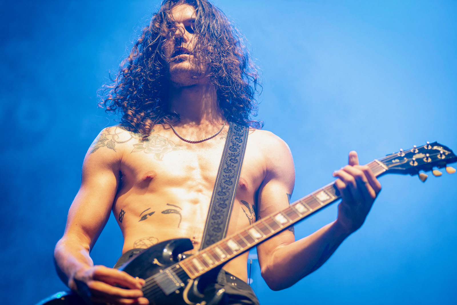 A shirtless Matty Bielawski plays a riff on his Gibson SG on a stage lit in blue.