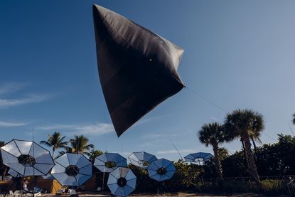Daytime image of Installation on miami beach, blue sky, palm trees, silver light reflectors, sand, floating grey inflated art piece object, held to the ground by rope cords
