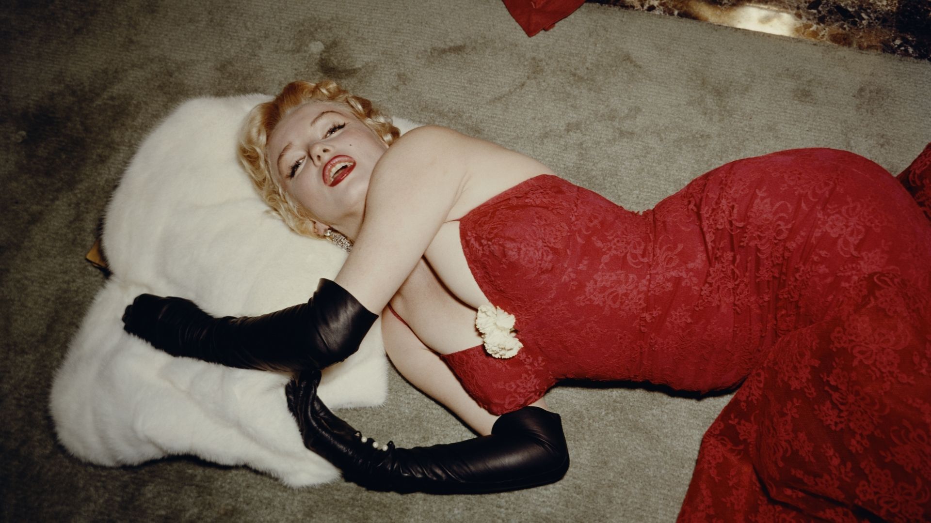 1955: EXCLUSIVE American actor Marilyn Monroe (1926 - 1962), wearing a red brocade evening gown and long black gloves, lying on a carpet and using a white fur stole as a pillow