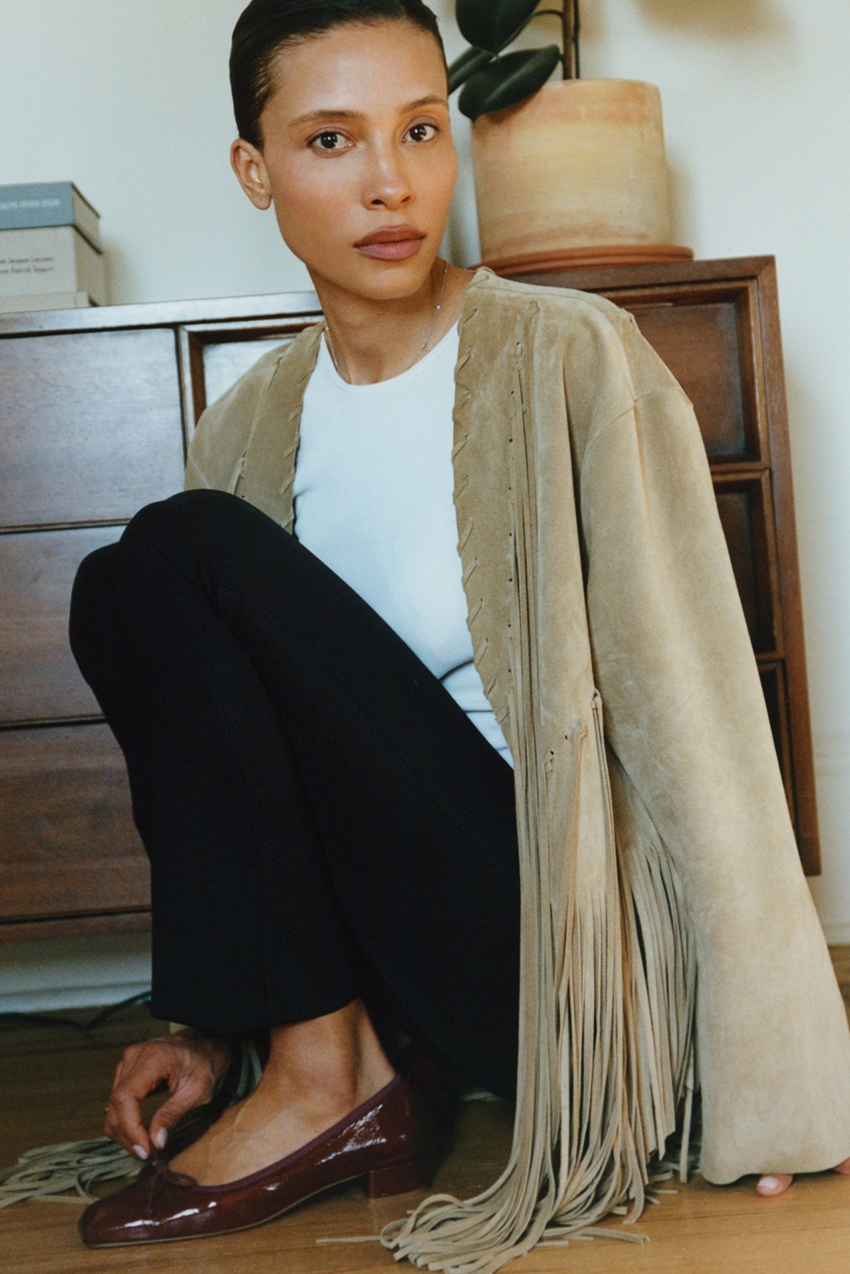 A woman wearing a tan jacket, white tee, black pants, and burgundy shoes.