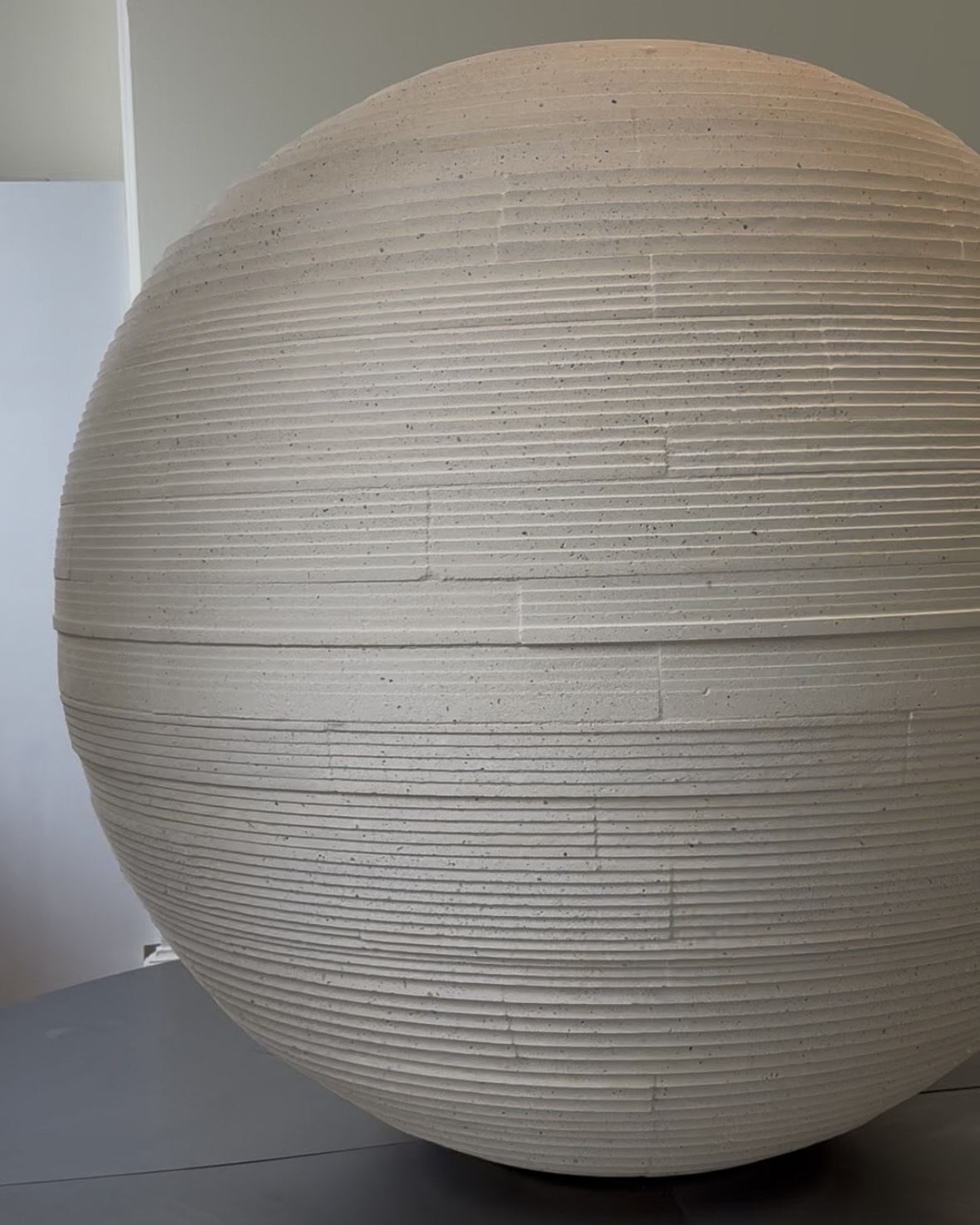 A brutalist installation made of a textural stone and comprising multiple linear sections superimposed to form a sphere sits in a naturally lit room.