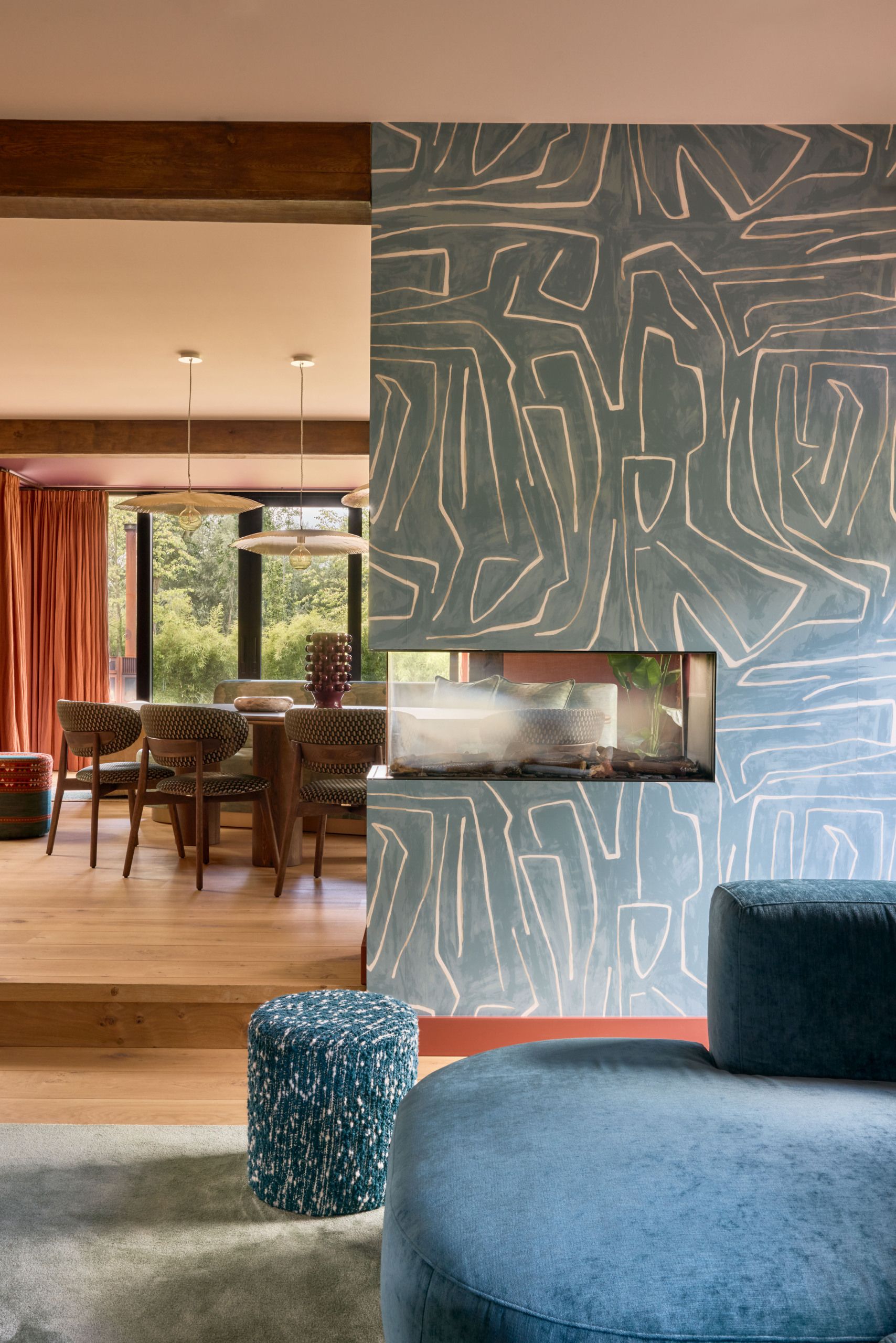 A living room that leads into a dining space with a half wall that has a printed blue wallpaper and a fire place in it.