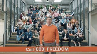 Man in orange sweater standing in front of people sitting in bleachers on Consumer Cellular commercial