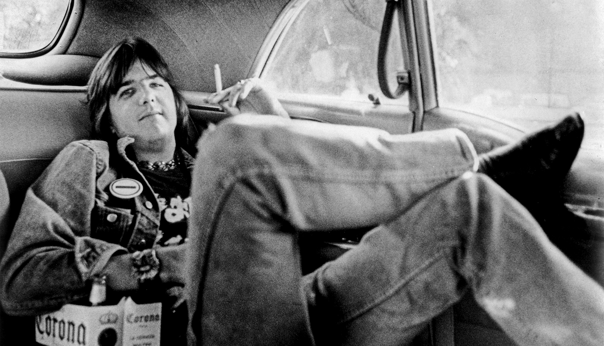 Singer/songwriter Gram Parsons poses for a portrait session wearing a t-shirt advertising his band Flying Burrito Bros. in the backseat of a Cadillac with a six pack of Corona beer in circa 1971 in Los Angeles, California.