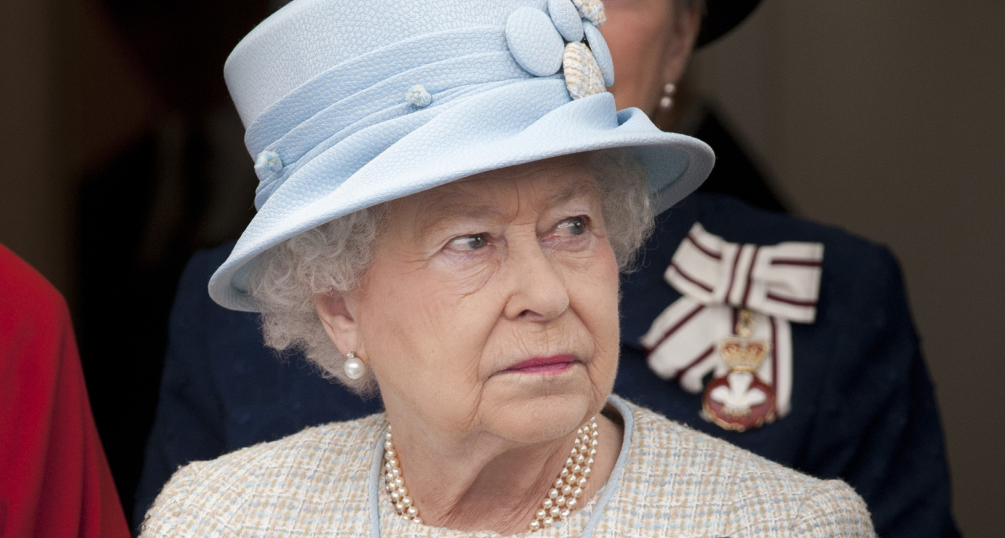 Queen Elizabeth wearing a blue hat and tweed coat with a serious expression