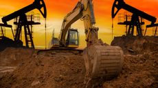 A backhoe flanked by oil rigs digs into the earth at sunset. 