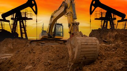 A backhoe flanked by oil rigs digs into the earth at sunset.