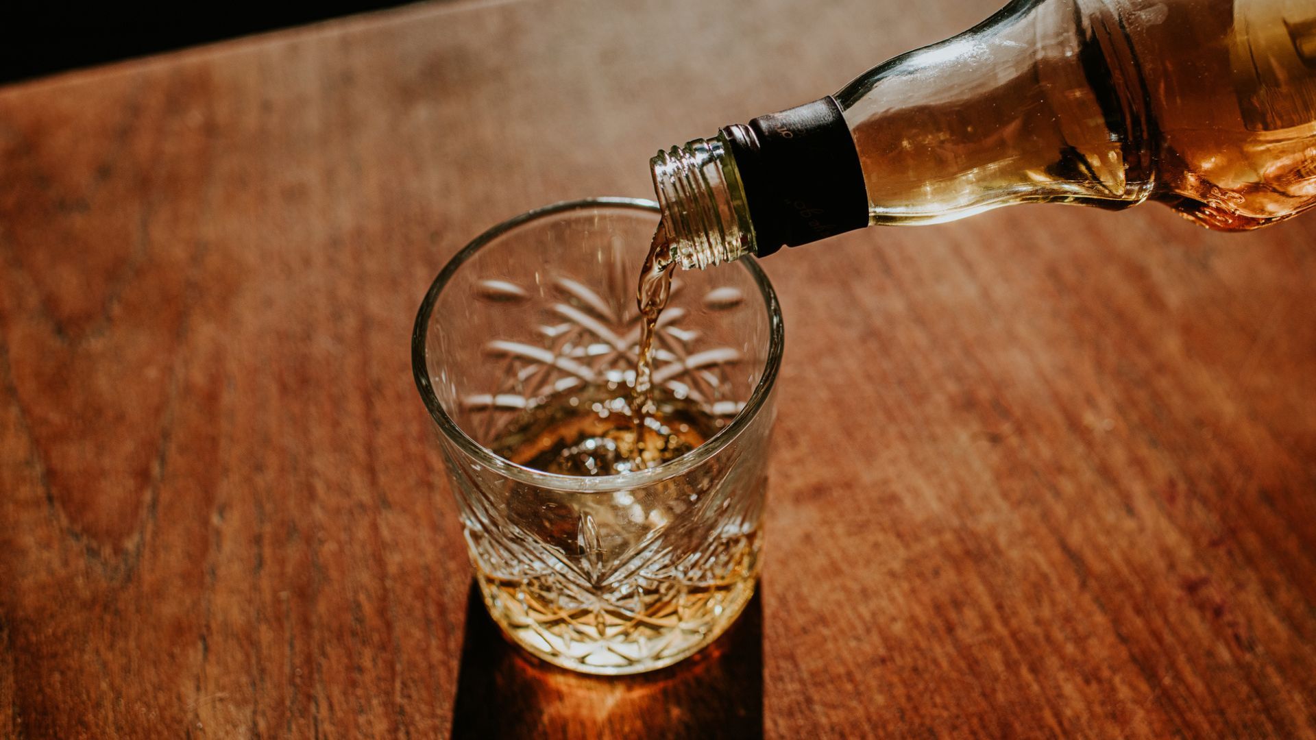 Person pouring whisky, one of the lowest calorie alcohols, into crystal bottom glass on wooden table