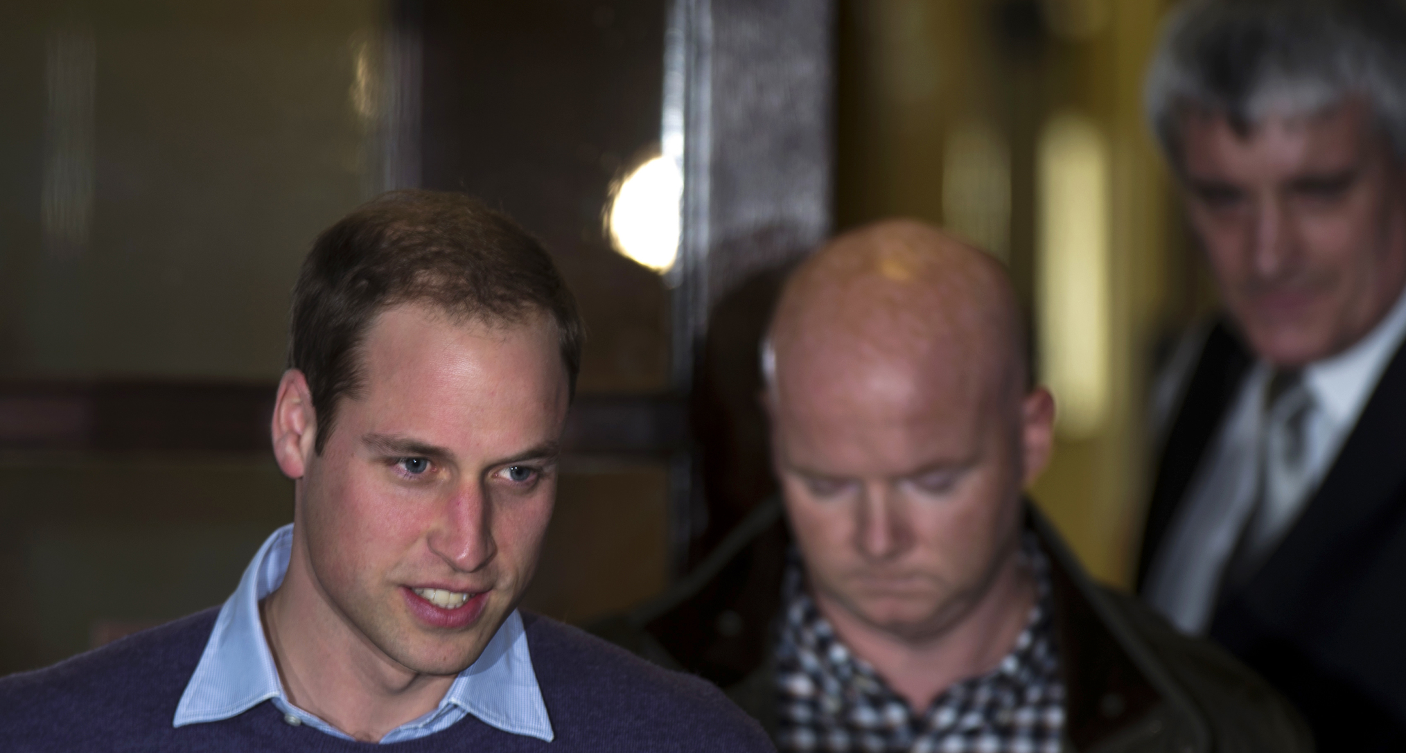 Prince William wearing a blue sweater with two men walking behind him