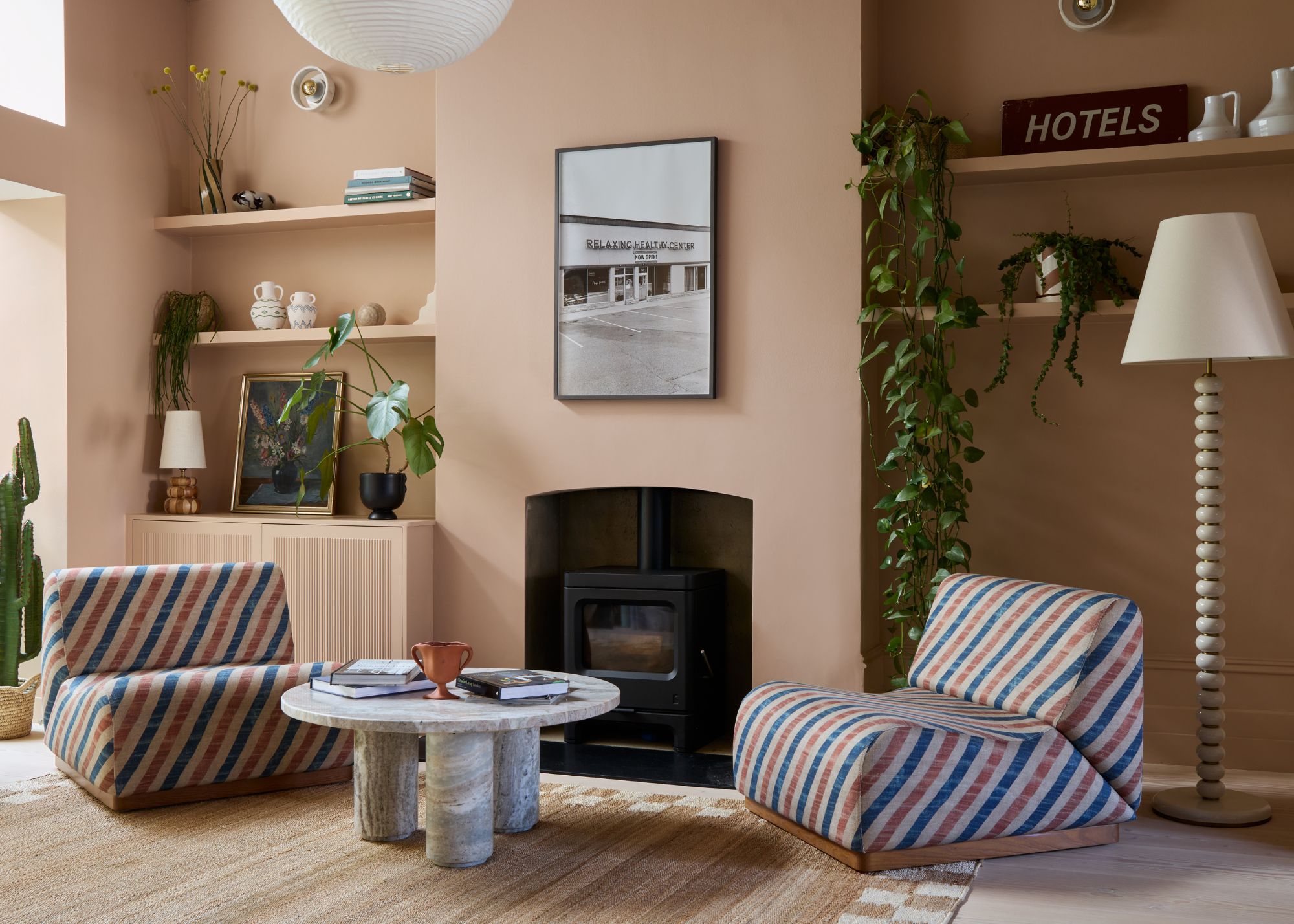 A living room color-drenched plaster pink, a log burner, two alcoves with open shelving and decorative objects, red and blue striped low-form chairs, wood floors, and a modern, round coffee table.