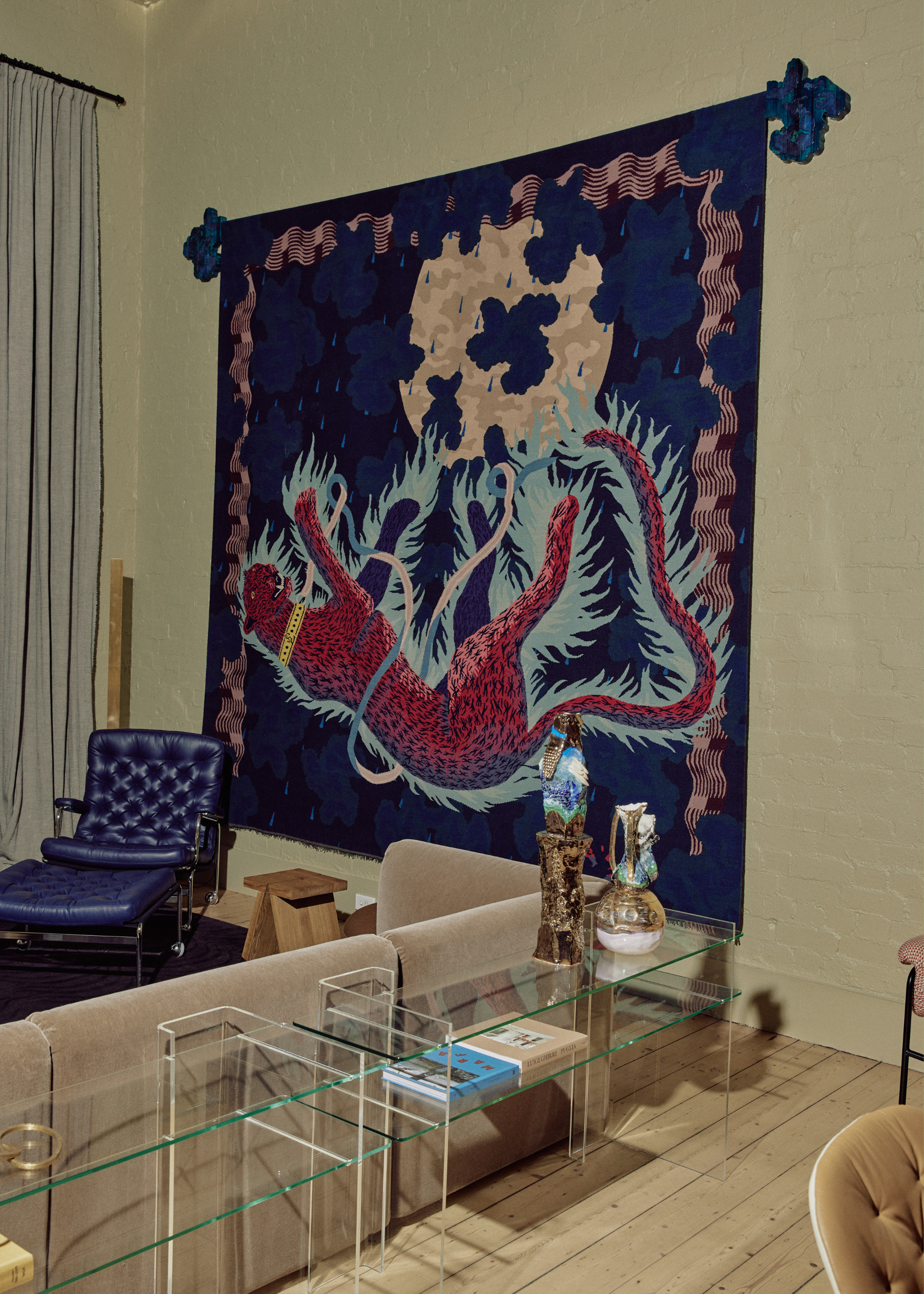 A living room with beige green walls, a large tapestry with a maroon leopard near a moon with blue clouds, a brown couch, a blue leather seat and a set of clear coffee tables with vases