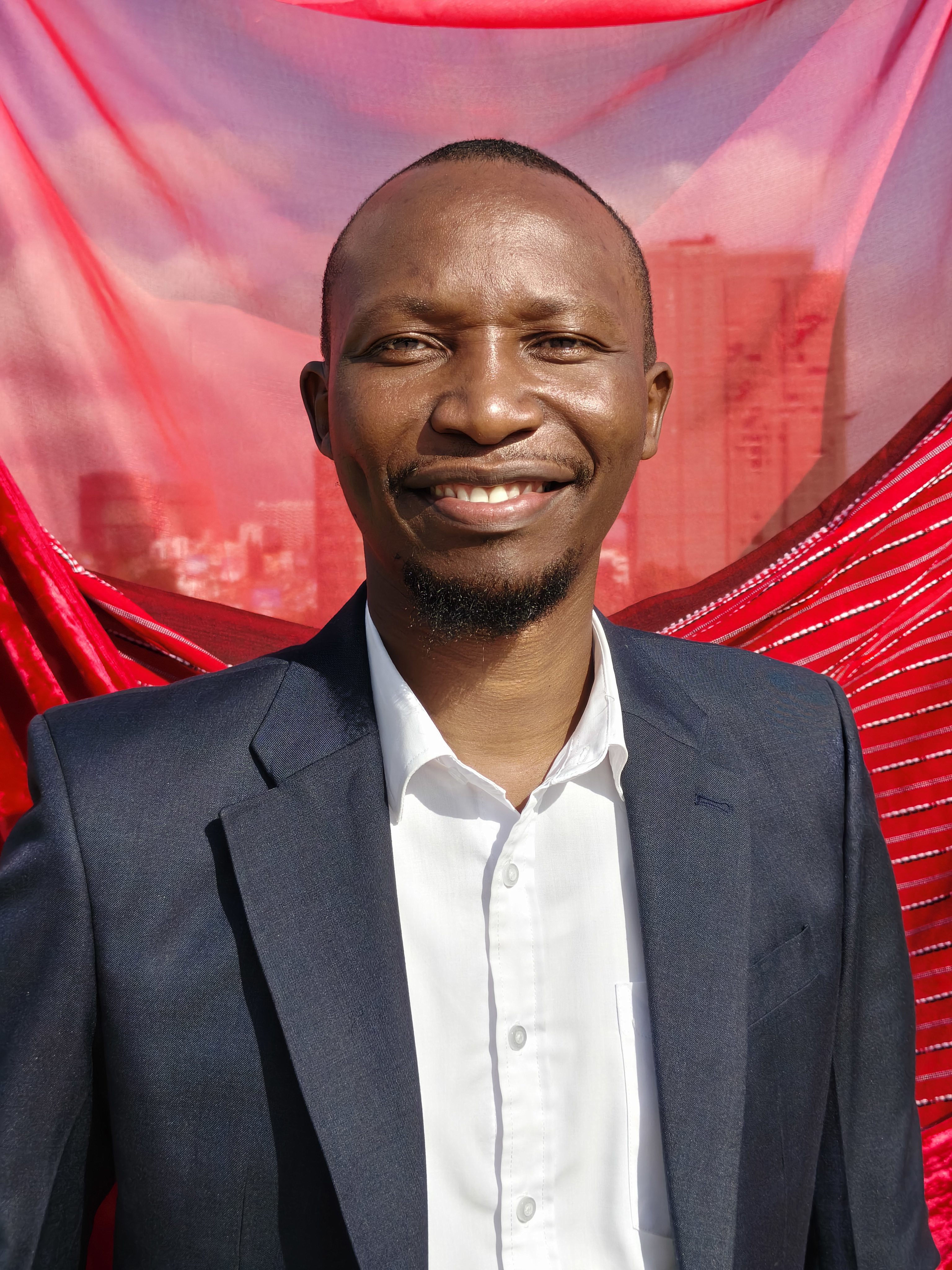 A smiling man in a navy blazer and open-collared white shirt poses in front of red striped fabric and a sheer red drape