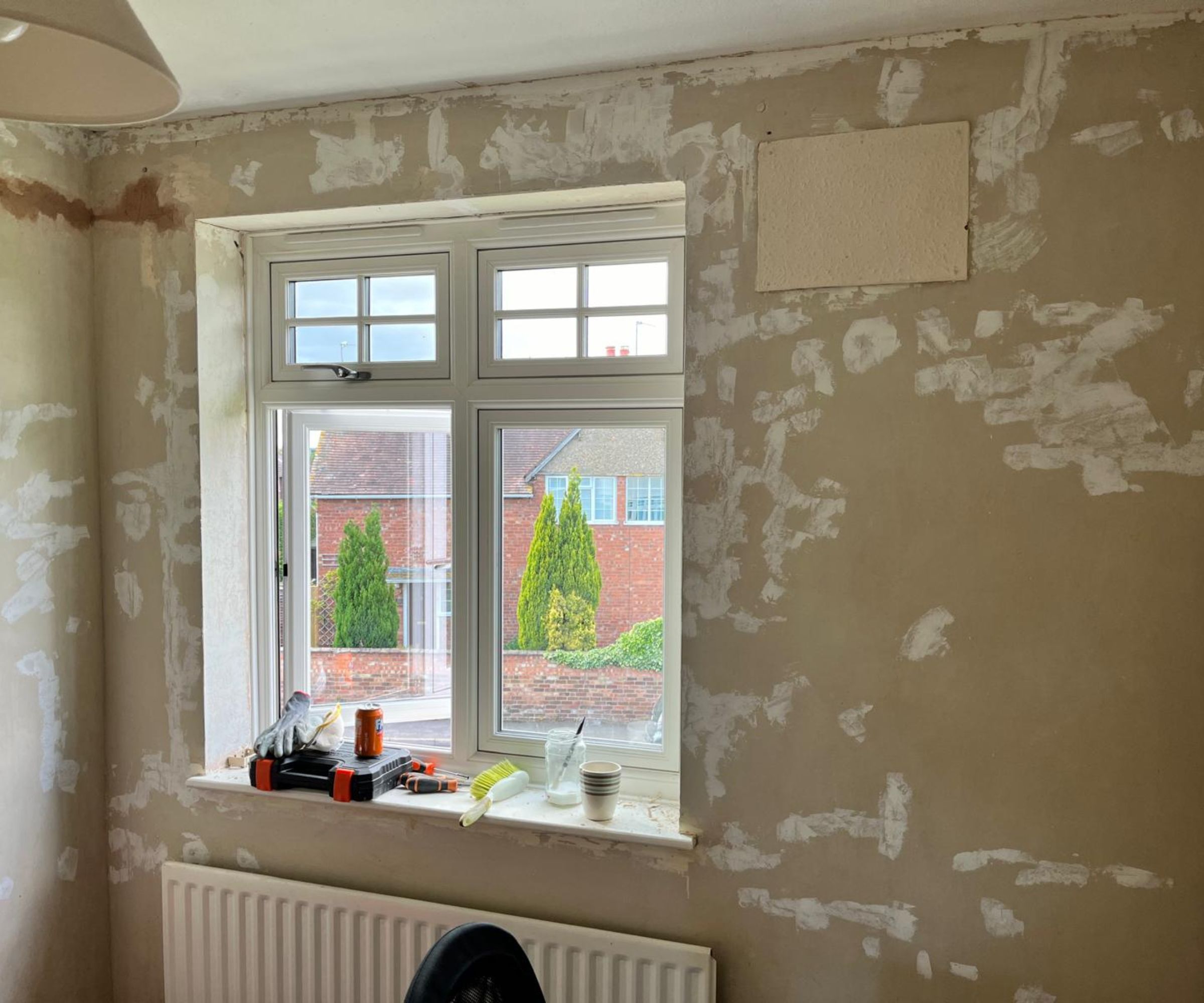 A room stripped back to bare plaster with patches of filler across the walls, a white uPVC window with tools on the sill, and a radiator below &amp;mdash; ready for replastering or painting.