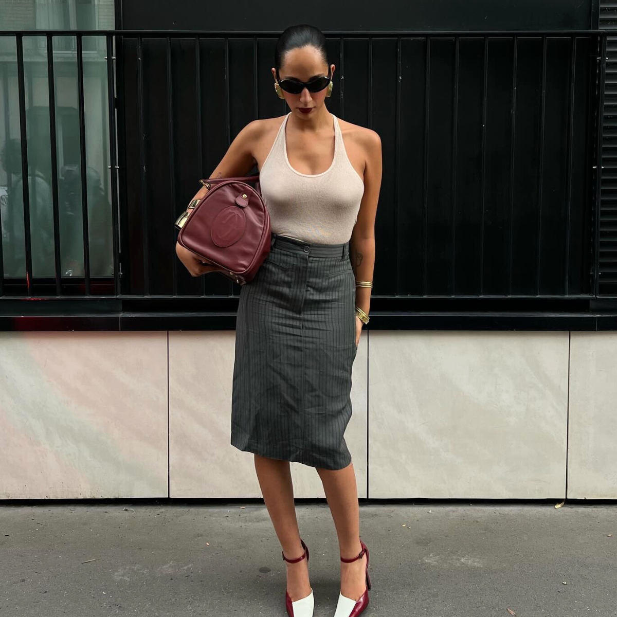 Fashion person Emily Nitta wears a cream halterneck top, a tailored skirt and two tone heel. She is carrying a matching burgundy duffle and oval sunglasses in a recent image from her Instagram. 