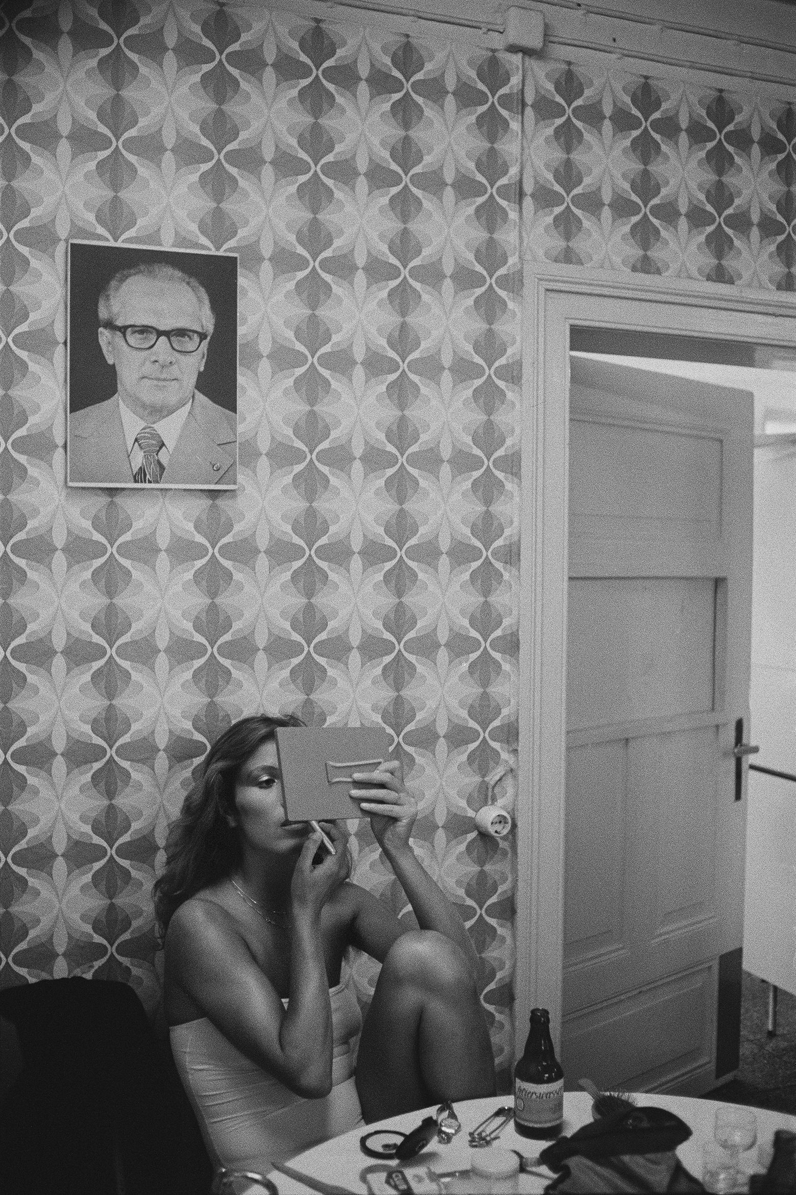 Black and white photo of a woman sitting at a table applying makeup in a vintage-patterned room with a portrait on the wall