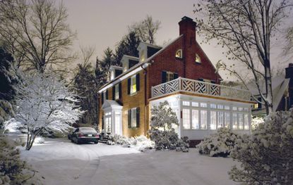 A colonial style home lit up from inside covered in snow outside, with a car out front.