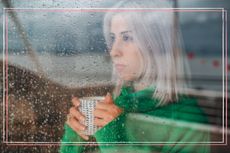 A woman looking out at rain through a window while holding a mug