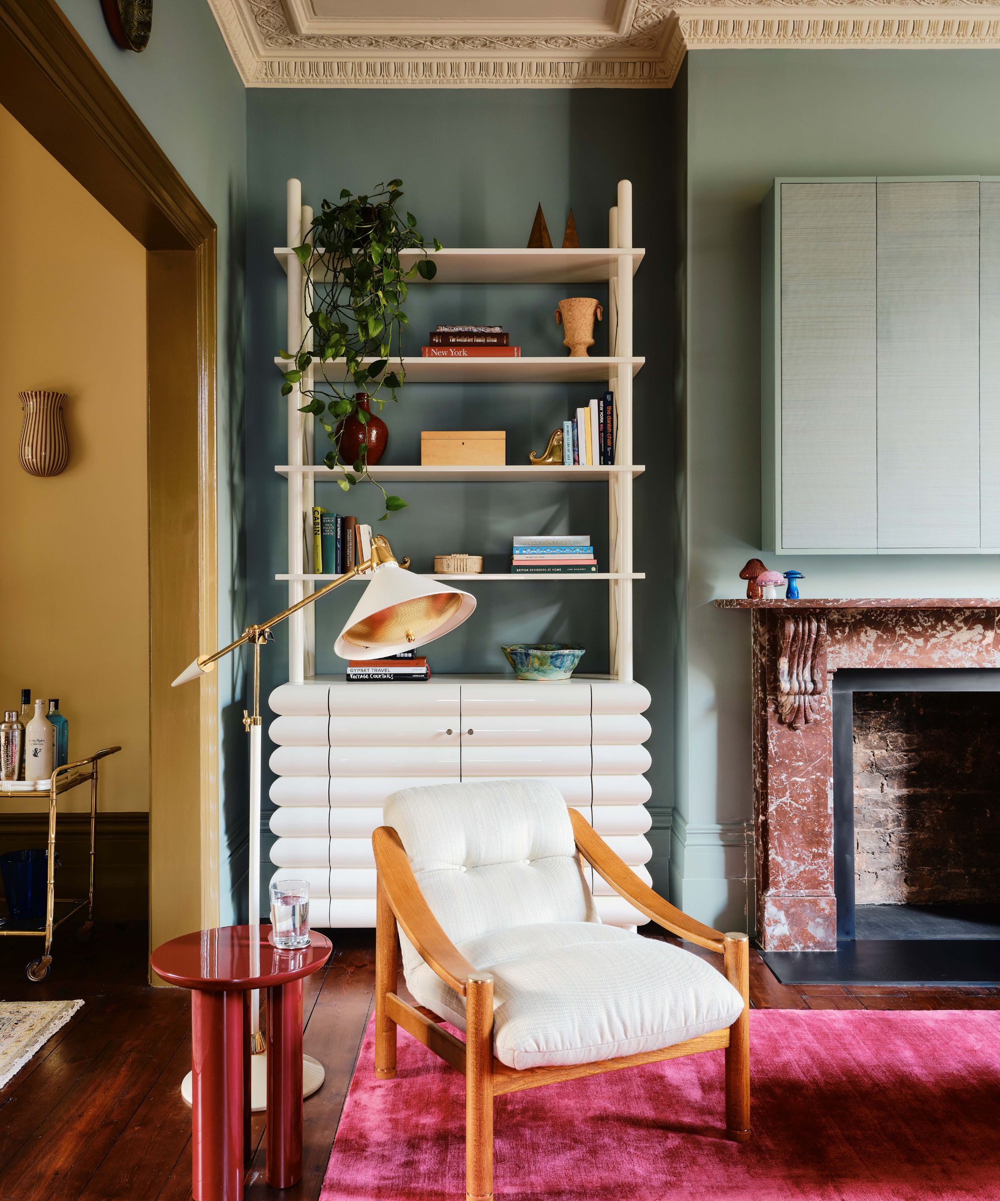 A living room with teal walls, a bright pink rug, a white armchair, and a white shelving unit with decorative objects and books.