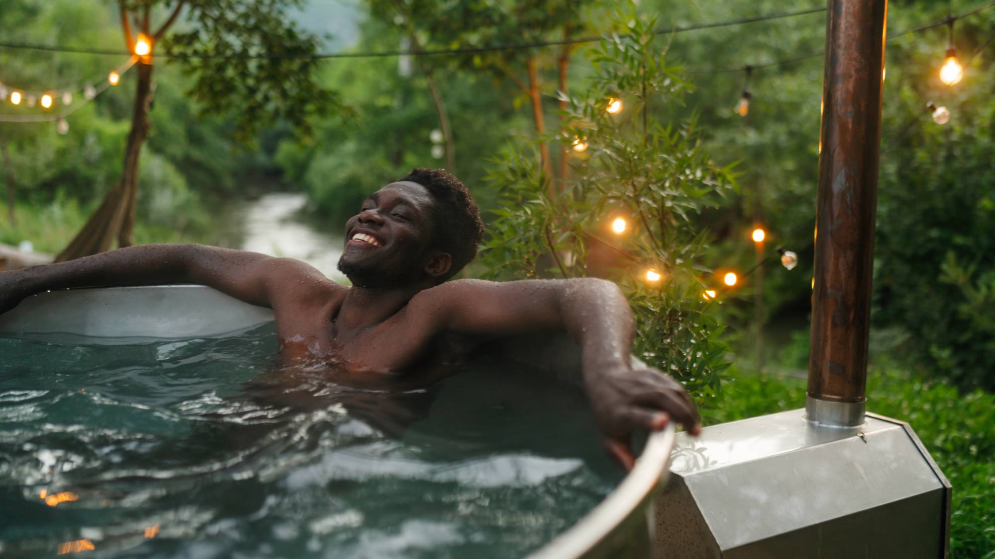 A smiling man relaxes in an outdoor tub surrounded by plants and string lights