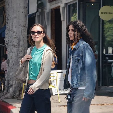 Lily Rose Depp wearing a teal shirt, beige hoodie, indigo jeans, suede shoulder bag, and tan suede loafers