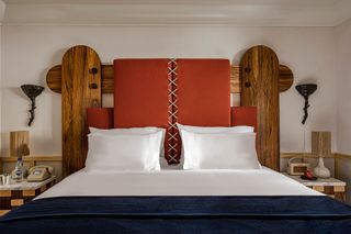 A wood and red leather sculptural bed board decorates a hotel room characterized by modern rustic furniture and whimsical touches, such as chequered brown and beige bedside tables.